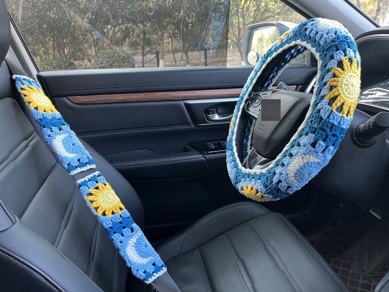 May include: A blue and yellow crocheted steering wheel cover with a sun and moon design. The cover is on a black leather steering wheel in a car.