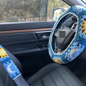 May include: A blue and yellow crocheted steering wheel cover with a sun design. The cover is on a black leather steering wheel in a car.