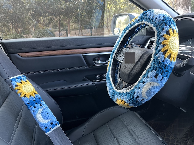 May include: A blue and yellow crocheted steering wheel cover with a sun design. The cover is on a black leather steering wheel in a car.