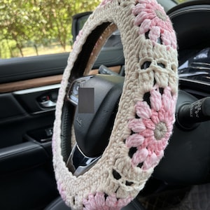 K&ouml;nnte beinhalten: Ein geh&auml;kelter Lenkradbezug mit rosa und beigefarbenen Blumen. Der Bezug ist aus weichem Garn gefertigt und soll Ihrem Auto Komfort und Stil verleihen.