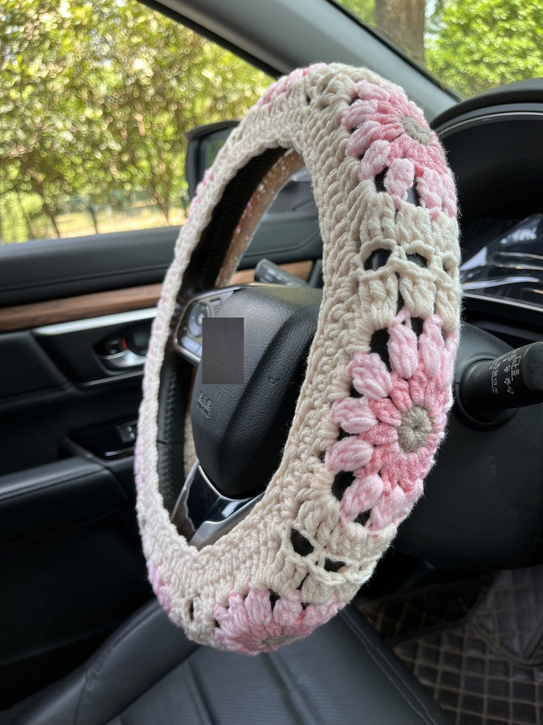 K&ouml;nnte beinhalten: Ein geh&auml;kelter Lenkradbezug mit rosa und beigefarbenen Blumen. Der Bezug ist aus weichem Garn gefertigt und soll Ihrem Auto Komfort und Stil verleihen.