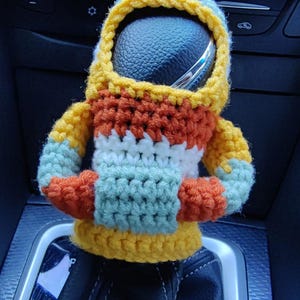 May include: A crocheted, multi-coloured, astronaut-shaped car gear shift knob cover. The cover is yellow, orange, white, and green.
