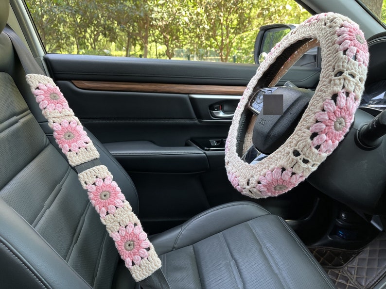K&ouml;nnte beinhalten: Ein Autoinnenraum mit einer geh&auml;kelten Lenkradabdeckung und Sicherheitsgurtbez&uuml;gen. Die Bez&uuml;ge sind rosa und beige mit einem Blumenmuster.