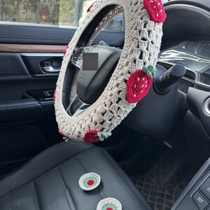 May include: A beige crocheted steering wheel cover with red and green strawberry accents. Two matching crocheted strawberry coasters are on the black leather seat.