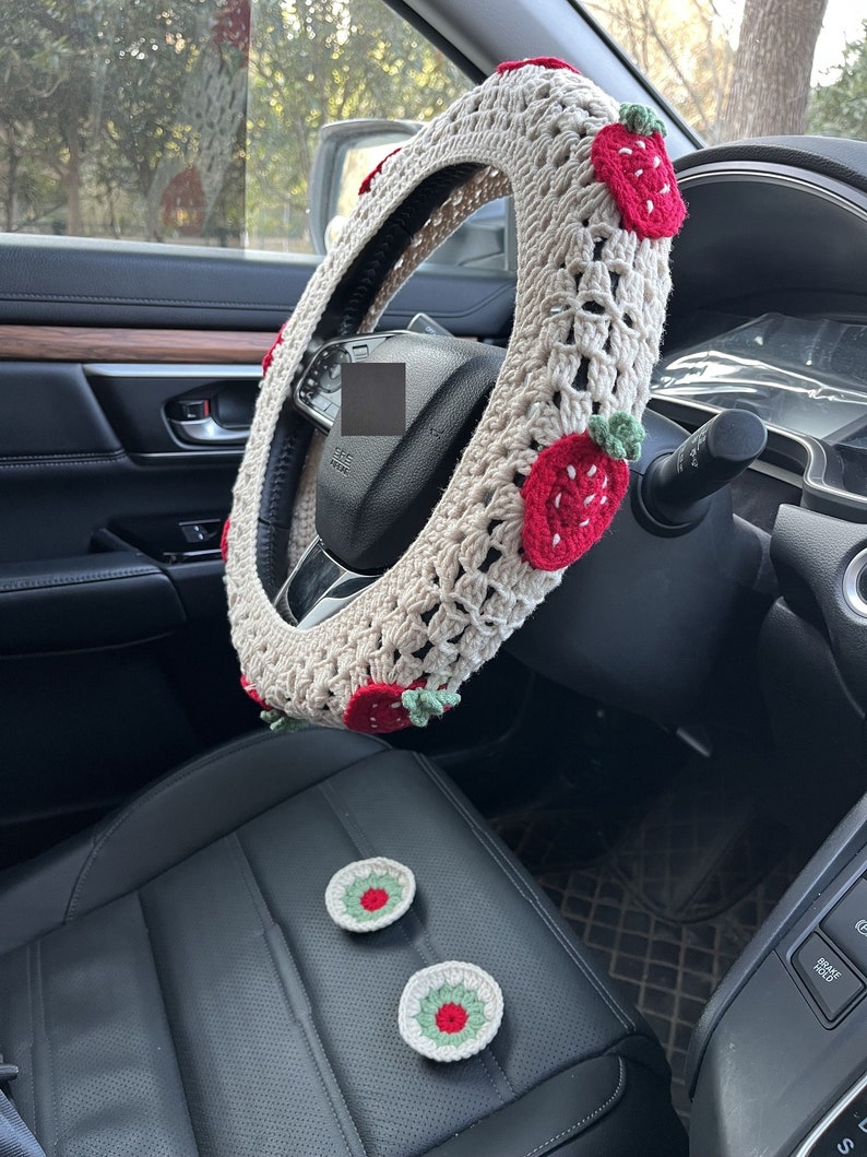 May include: A beige crocheted steering wheel cover with red and green strawberry accents. Two matching crocheted strawberry coasters are on the black leather seat.