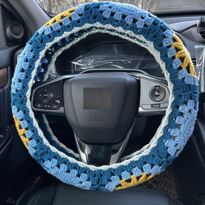 May include: A blue, white, and yellow crocheted steering wheel cover. The cover is made of a soft, textured yarn and has a unique, geometric pattern.