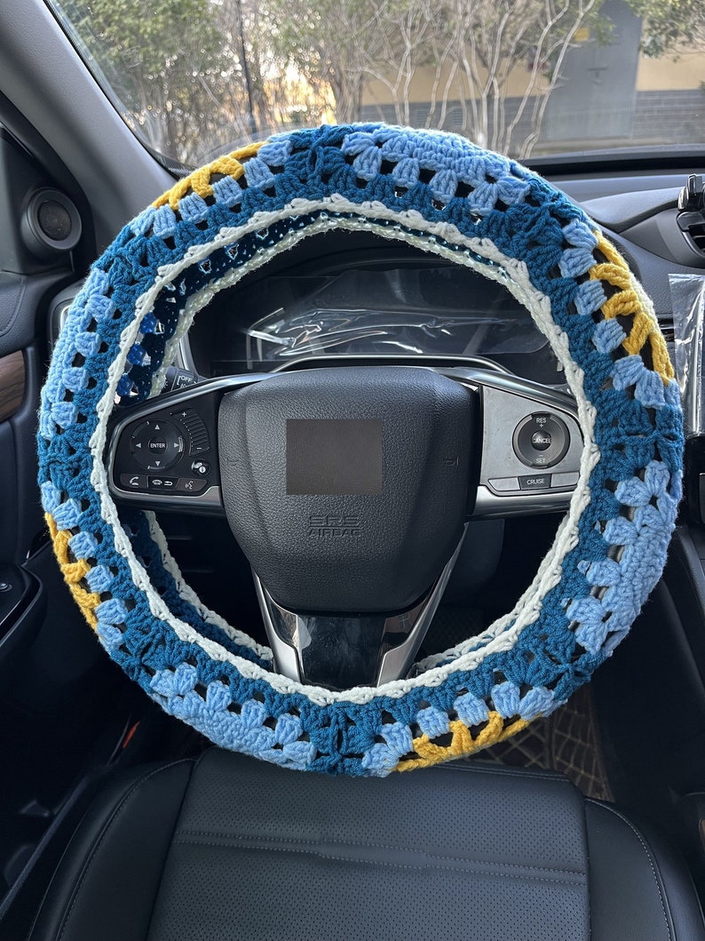 May include: A blue, white, and yellow crocheted steering wheel cover. The cover is made of a soft, textured yarn and has a unique, geometric pattern.