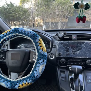 May include: A blue, white, and yellow crocheted steering wheel cover. The cover is on a black steering wheel in a car interior.