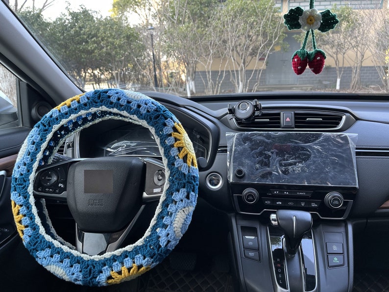 May include: A blue, white, and yellow crocheted steering wheel cover. The cover is on a black steering wheel in a car interior.