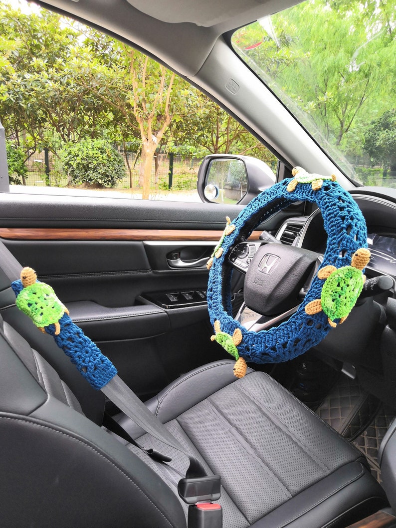 May include: A blue crocheted steering wheel cover with green crocheted turtles attached. The cover is fitted to a car's steering wheel. A green crocheted turtle is also attached to the car's seatbelt.