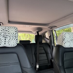 May include: Two white crocheted headrest covers for car seats. The covers are made with a square pattern and are placed on the headrests of the back seats of a car.