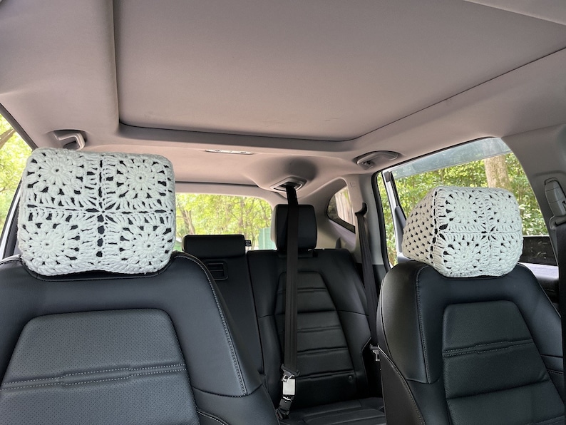 May include: Two white crocheted headrest covers for car seats. The covers are made with a square pattern and are placed on the headrests of the back seats of a car.