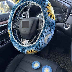 May include: A blue, yellow, and white crocheted steering wheel cover with two matching crocheted flower-shaped car accessories.