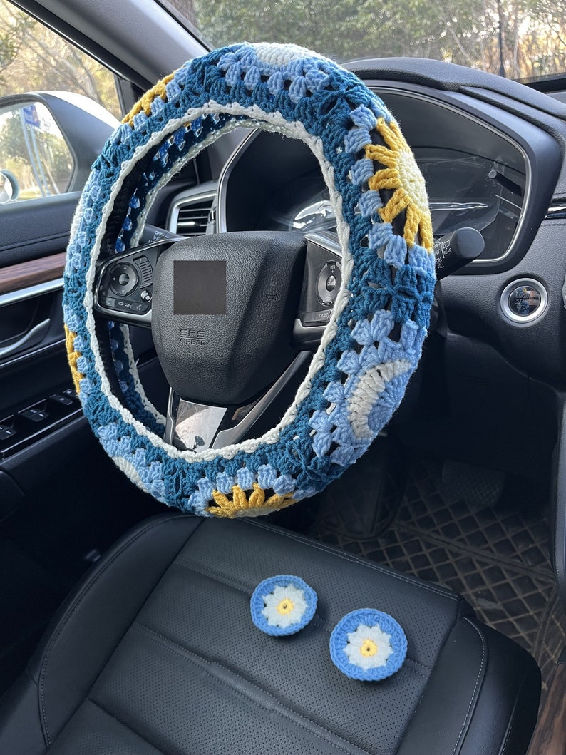 May include: A blue, yellow, and white crocheted steering wheel cover with two matching crocheted flower-shaped car accessories.