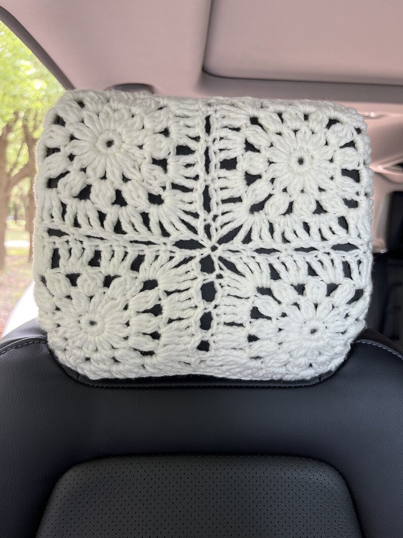 May include: White crocheted headrest cover with a square pattern. The cover is made of a soft, white yarn and has a delicate, floral design.