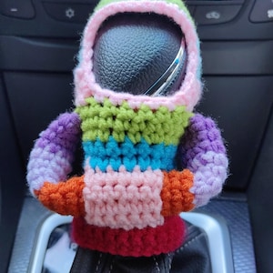 Peut inclure: Un couvre-levier de vitesses en crochet coloré en forme de personne portant un casque. Le couvre-levier est composé de rayures de différentes couleurs, notamment le vert, le violet, le bleu, le rose, l'orange et le rouge.