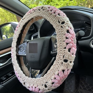 K&ouml;nnte beinhalten: Ein geh&auml;kelter Lenkradbezug mit beigefarbenem Hintergrund und rosa Blumenakzenten. Der Bezug ist aus Garn gefertigt und hat ein zartes, feminines Design.