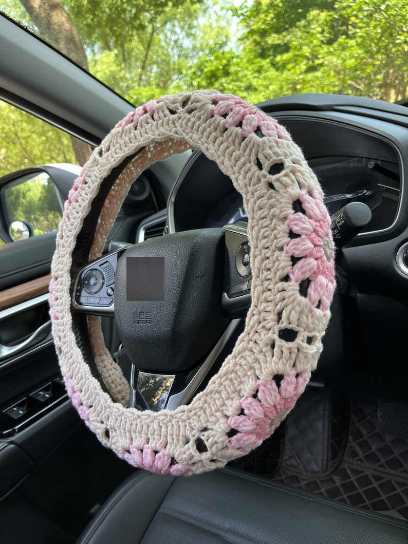 K&ouml;nnte beinhalten: Ein geh&auml;kelter Lenkradbezug mit beigefarbenem Hintergrund und rosa Blumenakzenten. Der Bezug ist aus Garn gefertigt und hat ein zartes, feminines Design.