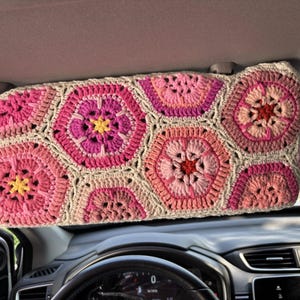 May include: A crochet sun visor cover with a floral design. The cover is made of pink, light pink, and beige yarn, with a pattern of hexagonal flower shapes. The visor is installed in a car.
