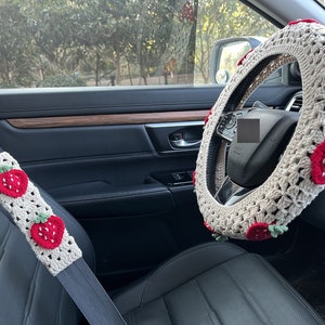 May include: A white crocheted steering wheel cover with red strawberry accents. The cover is installed on a car's steering wheel. A matching crocheted seatbelt cover with red strawberry accents is also visible.
