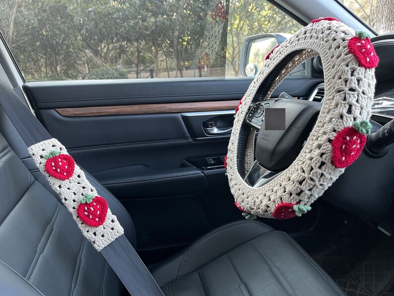 May include: A white crocheted steering wheel cover with red strawberry accents. The cover is installed on a car's steering wheel. A matching crocheted seatbelt cover with red strawberry accents is also visible.