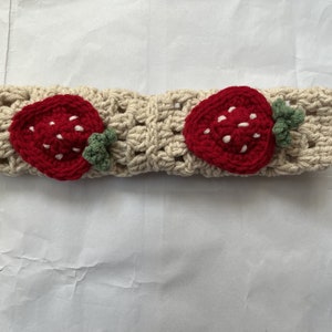May include: A crocheted headband with two red strawberry appliques. The headband is cream colored and the strawberries have white seeds.