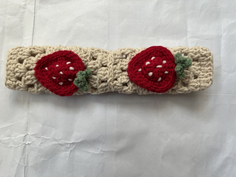 May include: A crocheted headband with two red strawberry appliques. The headband is cream colored and the strawberries have white seeds.