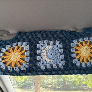 May include: A handmade, crocheted car sun visor cover. The cover is dark teal with three square panels. Each panel features a sun or moon design in yellow and light blue. The sun visor is installed in a car.