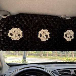 May include: A black, crocheted car visor featuring three white skull designs. The visor is rectangular and appears to be handmade, with a textured, open weave pattern. The skulls are centered and evenly spaced across the visor.