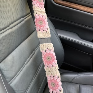 K&ouml;nnte beinhalten: Ein geh&auml;kelter Sicherheitsgurtbezug mit rosa und beigefarbenen Blumen. Der Bezug ist aus Garn gefertigt und hat ein geh&auml;keltes Design.