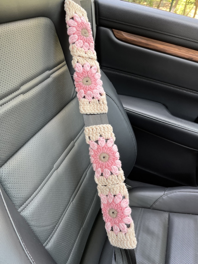 K&ouml;nnte beinhalten: Ein geh&auml;kelter Sicherheitsgurtbezug mit rosa und beigefarbenen Blumen. Der Bezug ist aus Garn gefertigt und hat ein geh&auml;keltes Design.