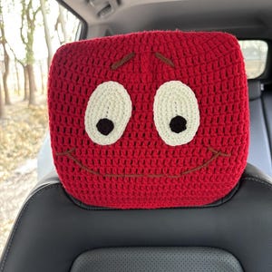 May include: Red crocheted car headrest with a smiling face. The headrest has white eyes with black pupils and a brown stitched smile.