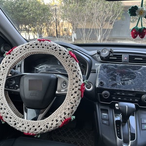 May include: A crocheted steering wheel cover with red and green accents. The cover is beige and has a delicate, intricate design. The cover is installed on a black steering wheel in a car.