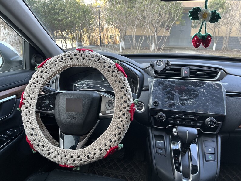 May include: A crocheted steering wheel cover with red and green accents. The cover is beige and has a delicate, intricate design. The cover is installed on a black steering wheel in a car.