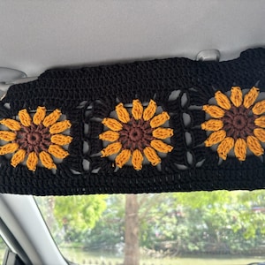 May include: Black crochet car visor featuring three sunflower designs. Each sunflower has a brown center and yellow petals. The visor is designed to attach to the car's existing visor.