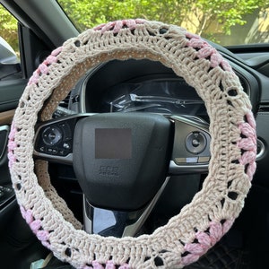 K&ouml;nnte beinhalten: Ein beigefarbener, geh&auml;kelter Lenkradbezug mit rosa Blumenakzenten. Der Bezug ist aus weichem Garn gefertigt und bietet einen komfortablen Griff.