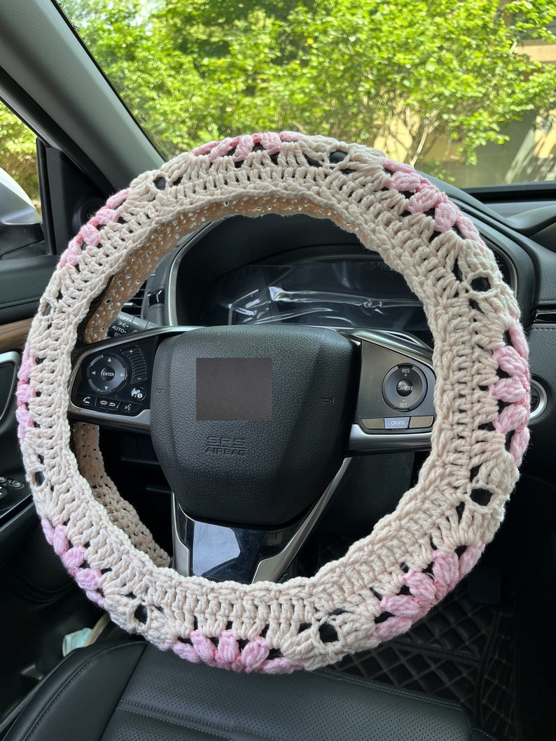 K&ouml;nnte beinhalten: Ein beigefarbener, geh&auml;kelter Lenkradbezug mit rosa Blumenakzenten. Der Bezug ist aus weichem Garn gefertigt und bietet einen komfortablen Griff.