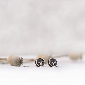 Puede incluir: Un par de pendientes de madera con un diseño de ciervo plateado. Los pendientes son redondos y tienen una veta de madera marrón oscura.