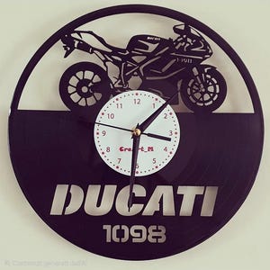 Può includere: Un orologio da parete circolare realizzato con un disco in vinile nero con la sagoma ritagliata di una moto Ducati 1098. Il quadrante bianco centrale mostra numeri neri, lancette nere e il testo rosso Cre_t_M sotto il punto centrale.