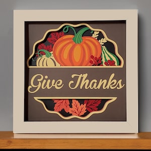 May include: A white framed shadow box with a brown background. The shadow box features a paper cut-out design of pumpkins, leaves, and berries. The text "Give Thanks" is written in a gold script font.