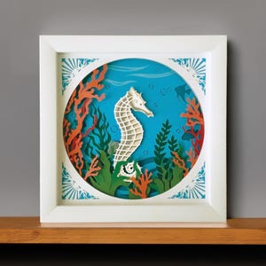 May include: A white paper seahorse with orange and green paper coral and seaweed in a blue paper ocean scene. The paper art is framed in a white frame.