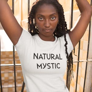 K&ouml;nnte beinhalten: Wei&szlig;es T-Shirt mit dem schwarzen Aufdruck "NATURAL MYSTIC". Das Model tr&auml;gt das T-Shirt und hat die Arme erhoben. Der Hintergrund umfasst ein Metalltor und eine gelbe Wand.