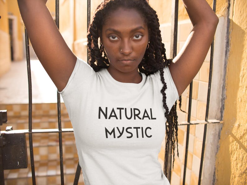 K&ouml;nnte beinhalten: Wei&szlig;es T-Shirt mit dem schwarzen Aufdruck "NATURAL MYSTIC". Das Model tr&auml;gt das T-Shirt und hat die Arme erhoben. Der Hintergrund umfasst ein Metalltor und eine gelbe Wand.