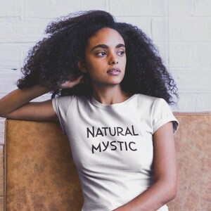 K&ouml;nnte beinhalten: Wei&szlig;es T-Shirt mit dem schwarzen Schriftzug "NATURAL MYSTIC". Die Person sitzt auf einem braunen Ledersofa, im Hintergrund eine wei&szlig;e Backsteinmauer.