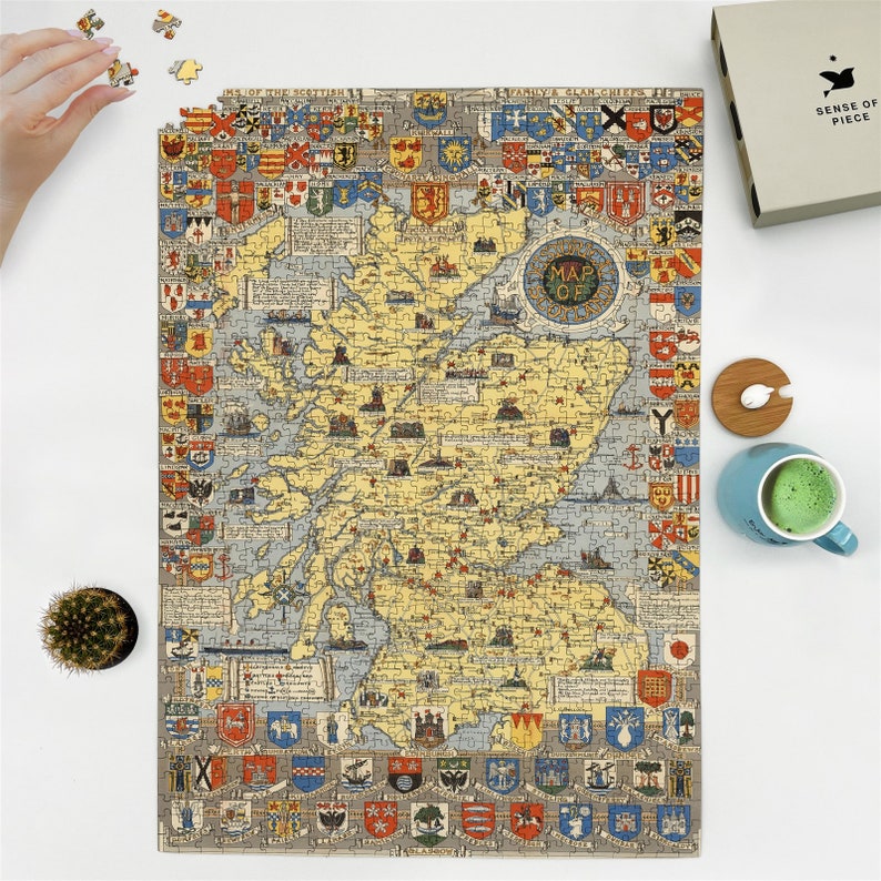 May include: A completed jigsaw puzzle depicting a vintage map of Scotland, featuring detailed illustrations and heraldic crests. The puzzle is mostly yellow and blue, with a circular "Map of" emblem. A hand is placing a puzzle piece.