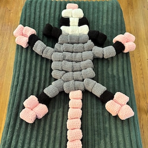 May include: A handmade, plush creature in a lizard-like form, crafted from soft, textured yarn. The body is primarily gray, with black and pink accents on the limbs and head. The creature is lying on a green, ribbed surface.