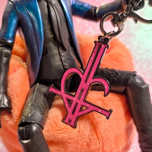 May include: A figurine of a character in a blue jacket and black outfit, seated on an orange cushion, holding a pink and black keychain. The keychain features a stylized letter design. The background is a sparkly pink.