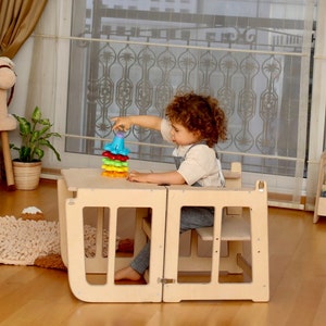 May include: A child sits at a wooden table and chair set, playing with a colorful stacking toy. The table and chair are designed for toddlers and have a safety gate around the seat.