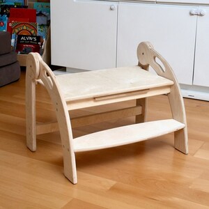 May include: A wooden step stool with a built-in table. The stool is made of light wood and has a curved design. The table is also made of light wood and has a small drawer.