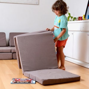 May include: A child is assembling a grey modular sofa. The sofa is made up of several sections that can be configured in various ways. The child is wearing a light blue shirt and red shorts.
